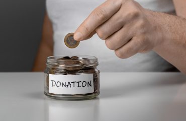 glass-money-jar-with-label-with-word-donations-hand-putting-penny-money-jar-charity-donation-cropped-view-man-putting-coin-penny-jar-with-donate-lettering